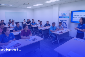 A photograph of a food safety training seminar for Filipino participants, conducted by Leadsmart PH. A female trainer presents at the front of a classroom, and multiple attendees are seated, observing the presentation on a screen and pull-up banners that read 'CERTIFIED FOOD SAFETY TRAINING'.