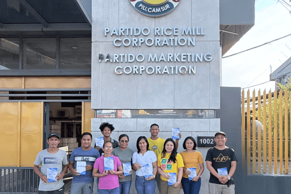 A group of food safety training participants posing for a group photo in front of the Partido Rice Mill Corporation logo during the LeadSmart PH Food Safety Compliance Officer training in Camarines Sur.
