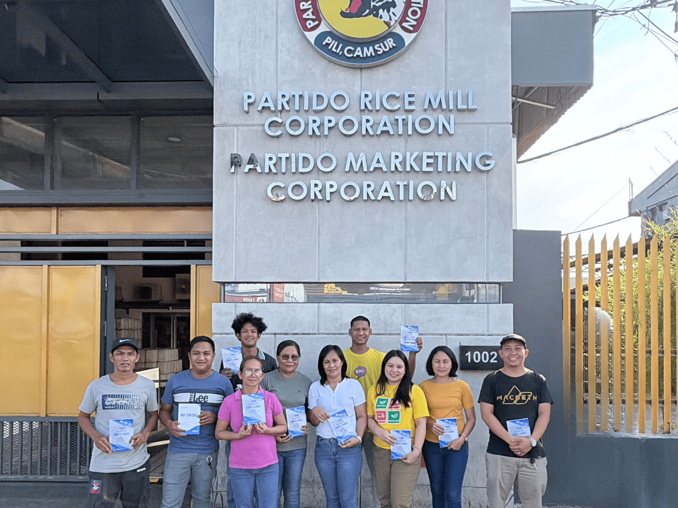 A group of food safety training participants posing for a group photo in front of the Partido Rice Mill Corporation logo during the LeadSmart PH Food Safety Compliance Officer training in Camarines Sur.