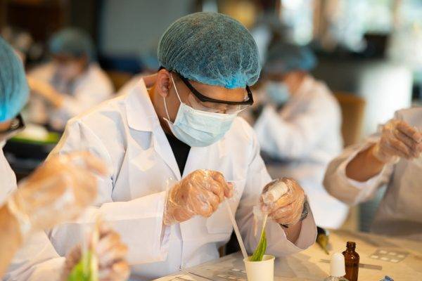 A food safety professional in a white lab coat, hairnet, and gloves performing a laboratory analysis on food samples, using a pipette and test tubes in a clean, brightly lit testing environment